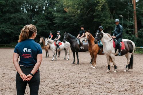 Horses and riders lined up on the centre line in an arena with the coach looking at them